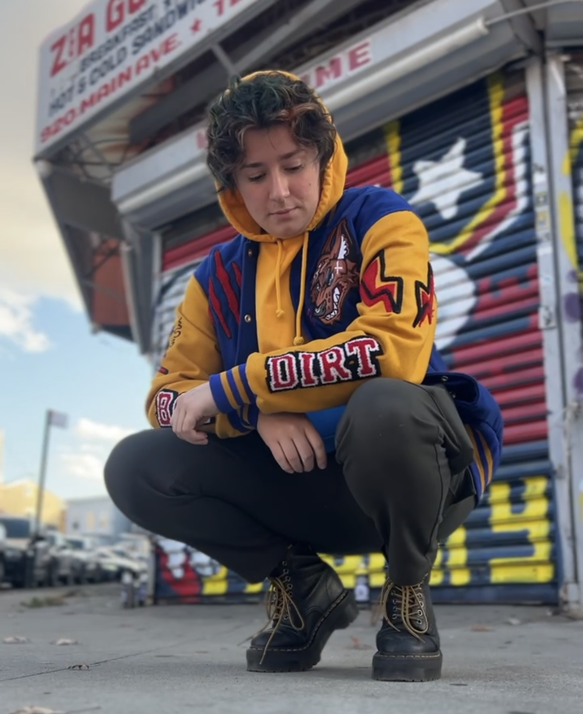 Person wearing a colorful jacket with 'Dirt' on it, squatting in front of a closed store.